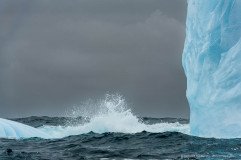 Vertical face of blue iceberg with breaking wave and dark clouds